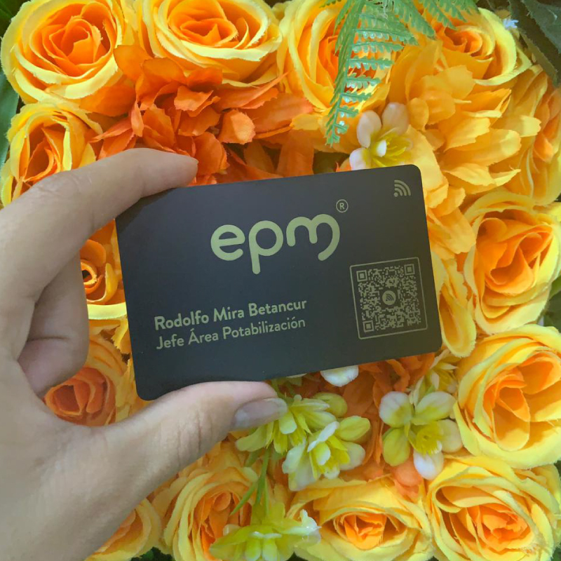 Black EPM business card held by a hand, displaying the name Rodolfo Mira Betancur and the title Jefe Área Potabilización, with a QR code among orange roses in the background.