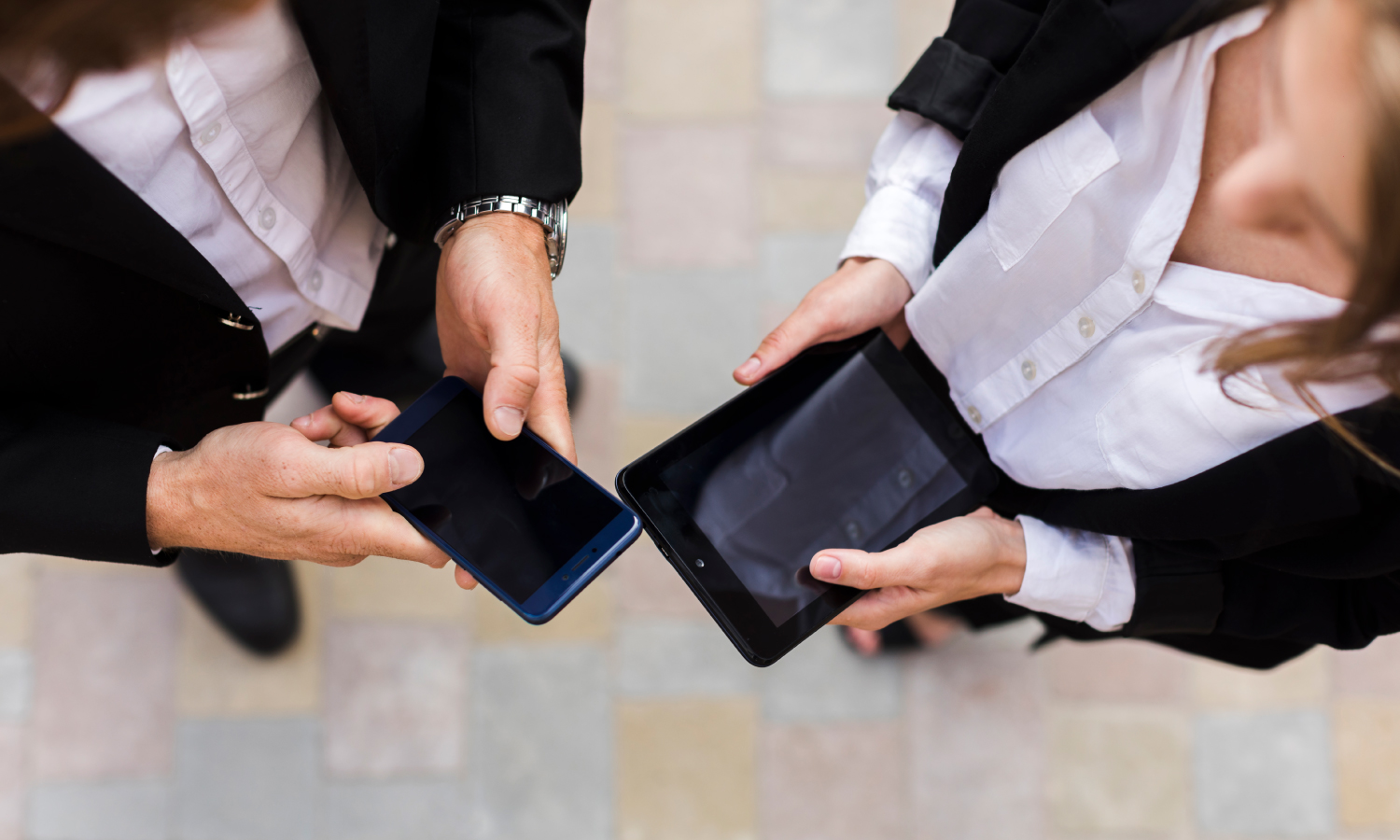 Two professionals in business attire hold a smartphone and a tablet, sharing the screen between them from above.