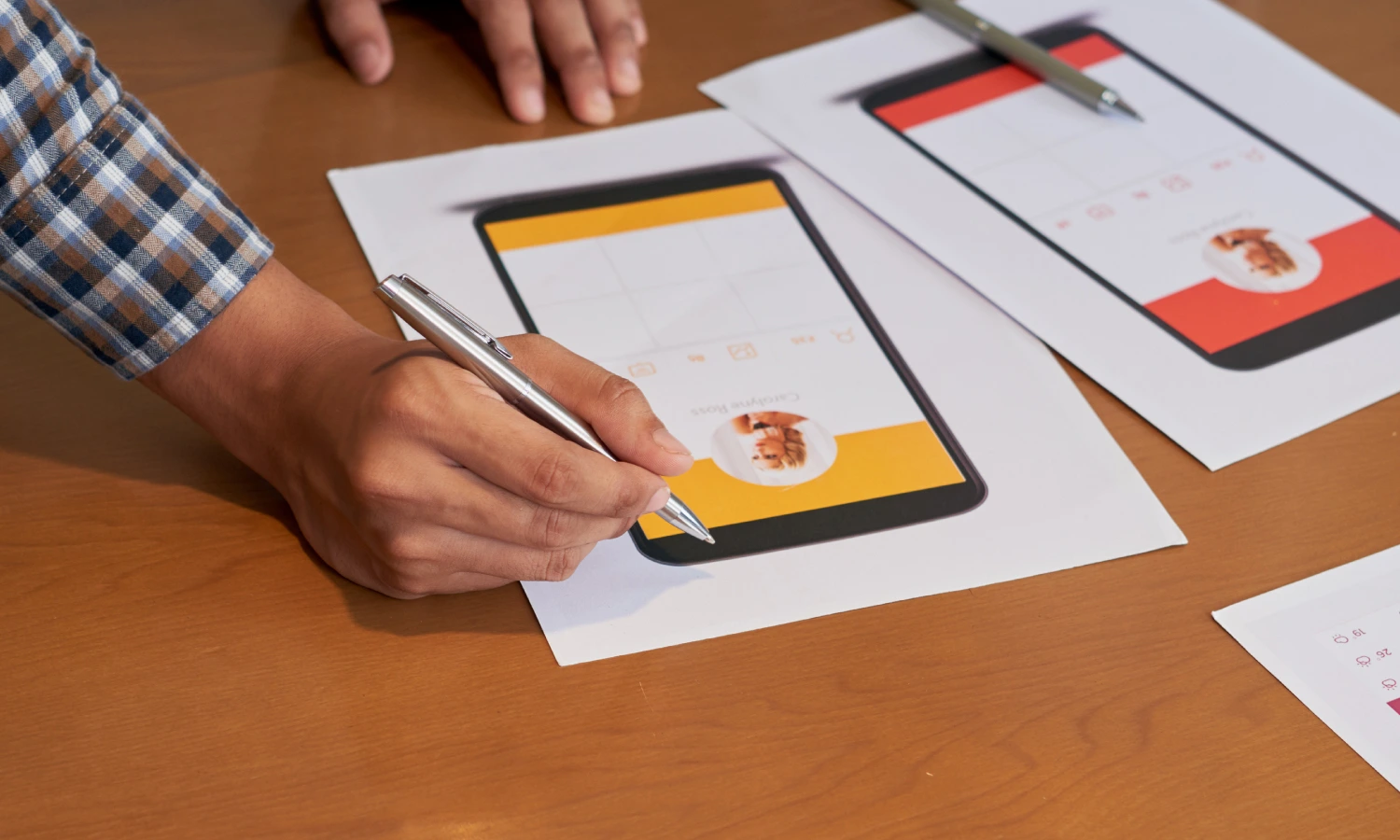 Designer in a plaid shirt sketching mobile app screens on printed mockups with a silver pen.