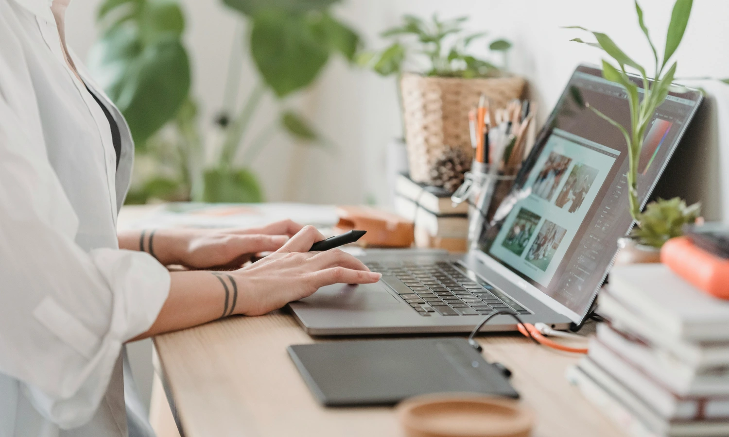 Person typing on a laptop at a wooden desk with a drawing tablet nearby, surrounded by plants and office supplies.