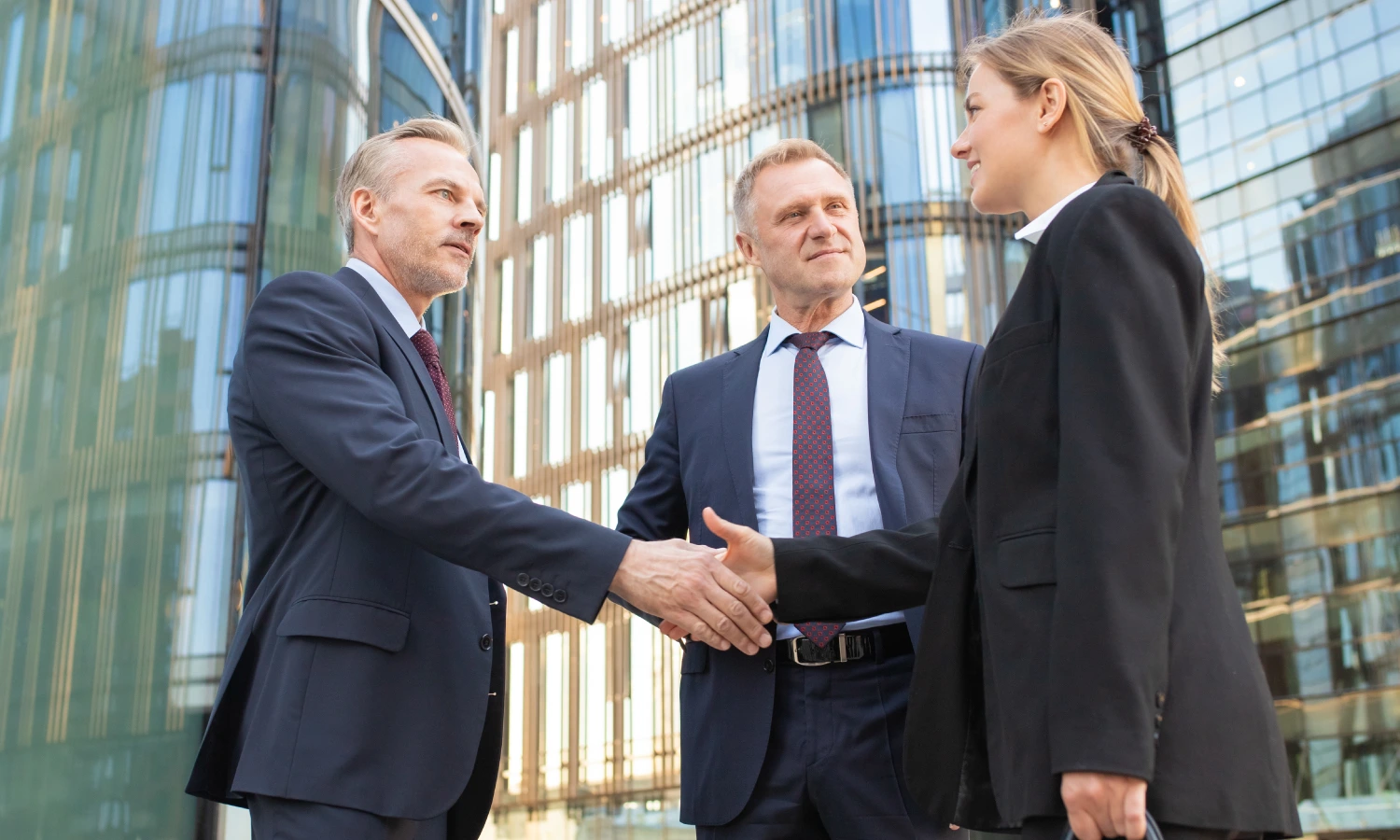 Two suited colleagues shake hands outside a glass-fronted office building, facing each other with a woman nearby.