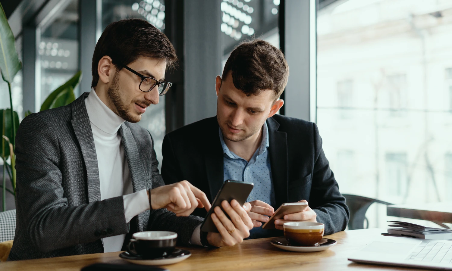 Two businessmen in blazers look at a smartphone together at a cafe, with one tapping the screen while the other holds a second device.