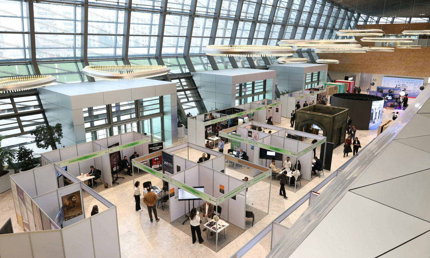 Bird's-eye view of a modern exhibition hall with white booth walls, attendees at tables, and circular ceiling lights above.