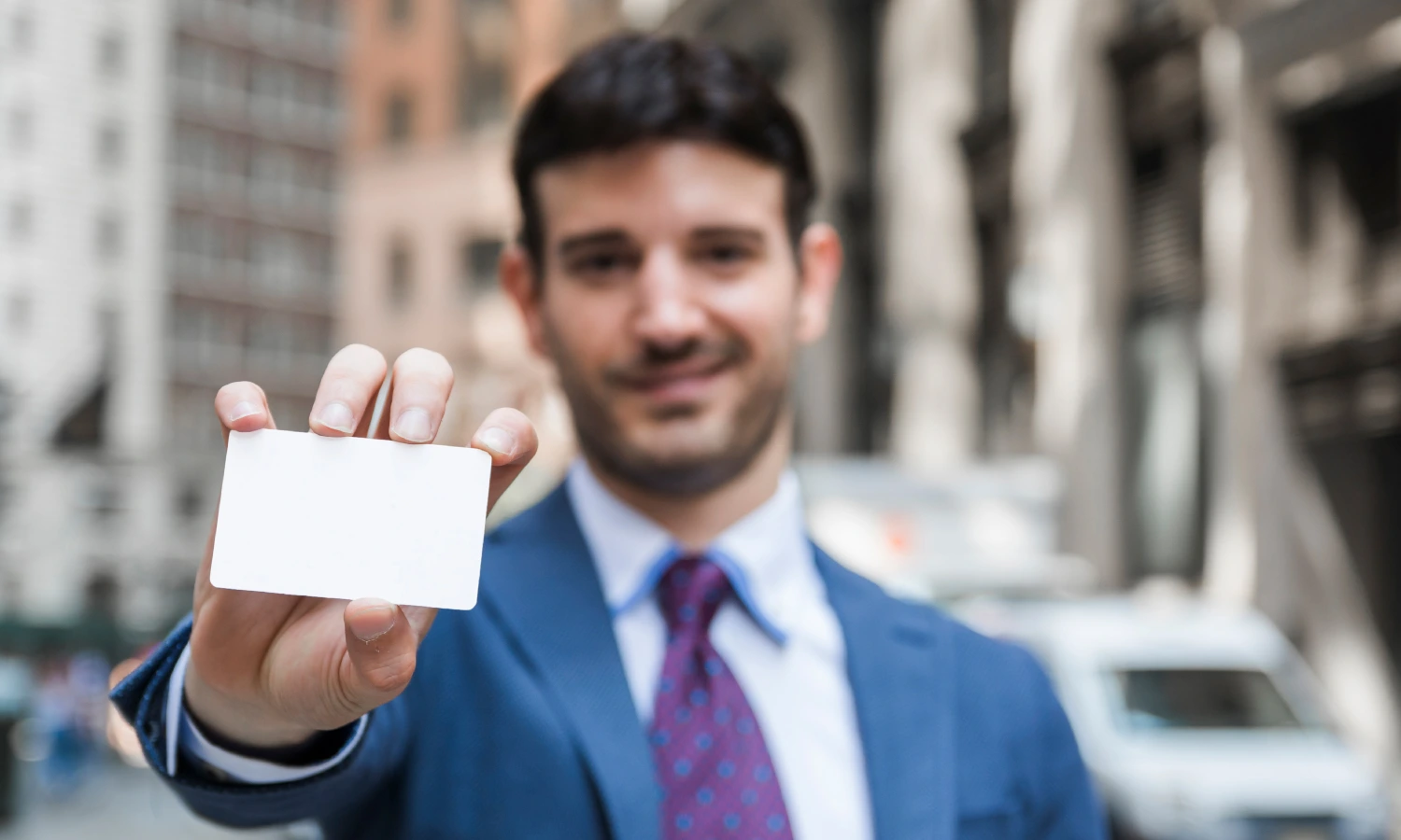 Businessman in a blue suit holding out a blank business card toward the camera on a city street.