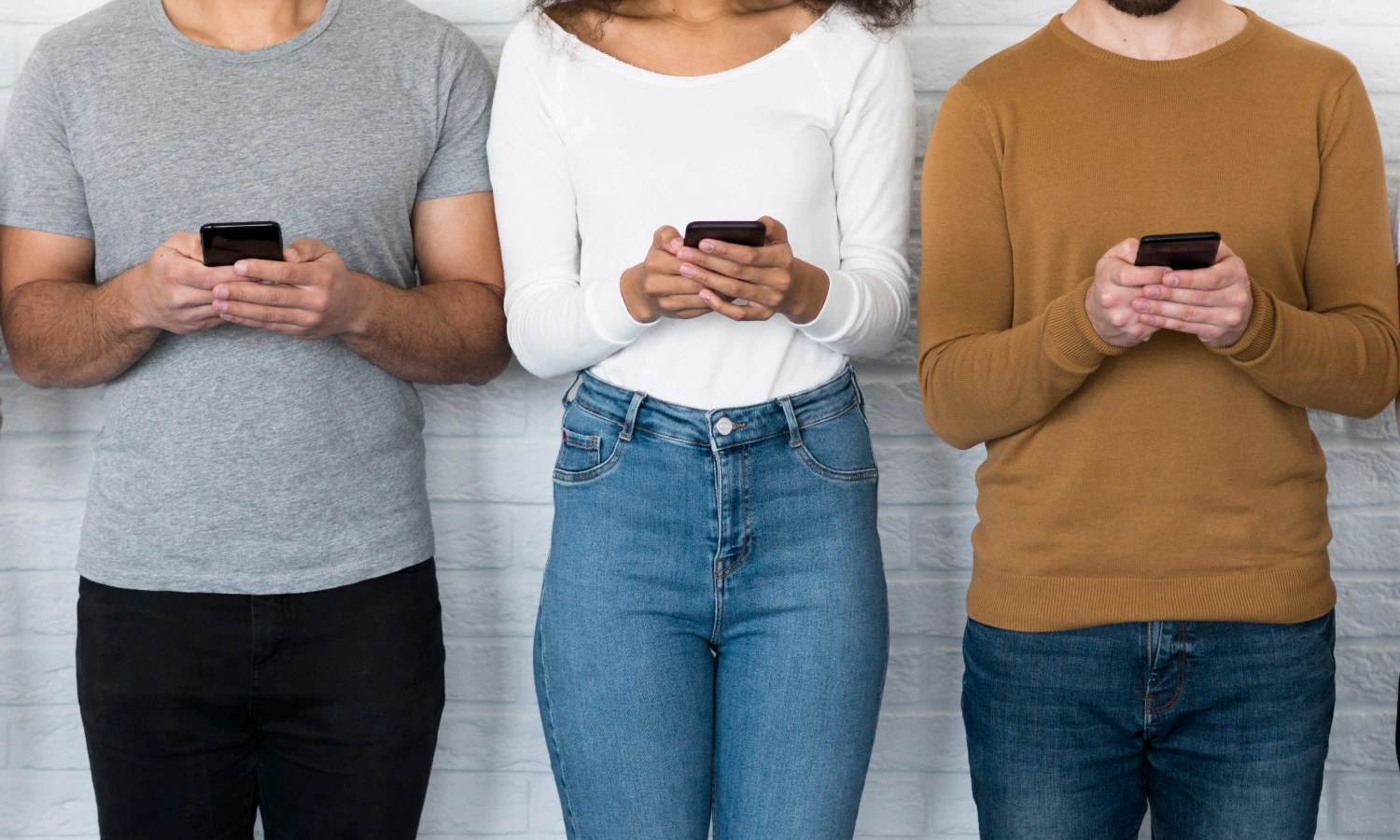 Three people standing against a light brick wall, each looking at a smartphone.