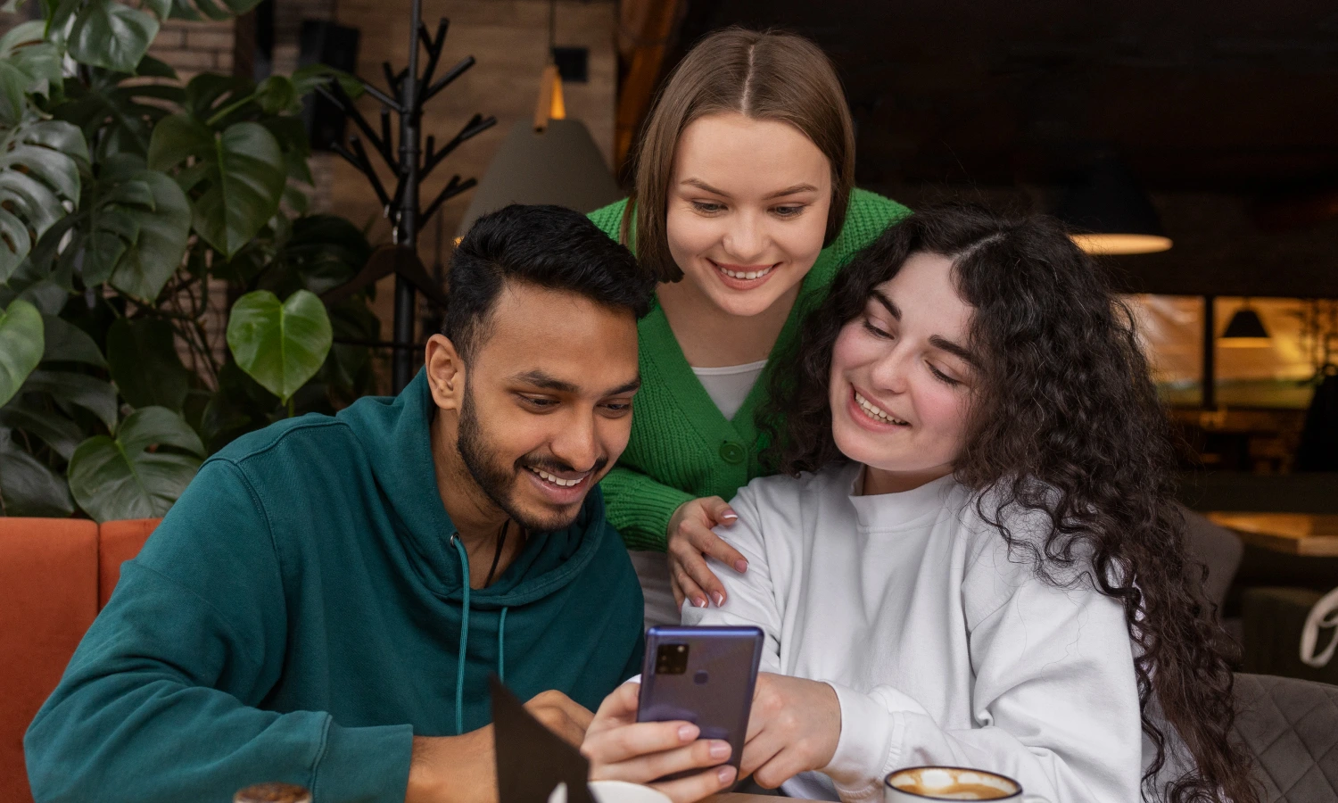 Three friends in a cafe huddle around a smartphone, smiling at the screen.