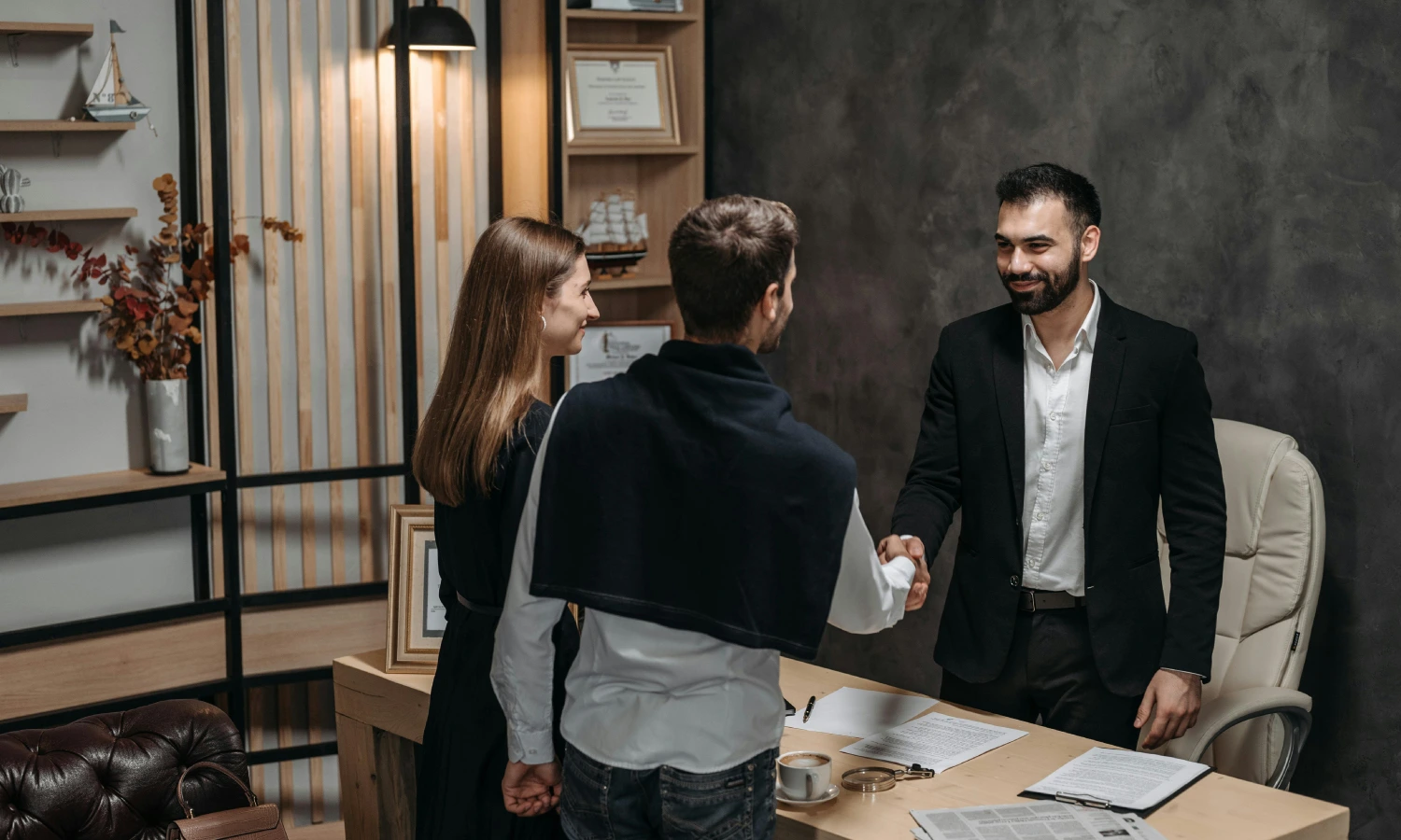 Man in a suit shakes hands with another man across a desk in a modern office while a woman watches, papers and coffee on the desk behind them.