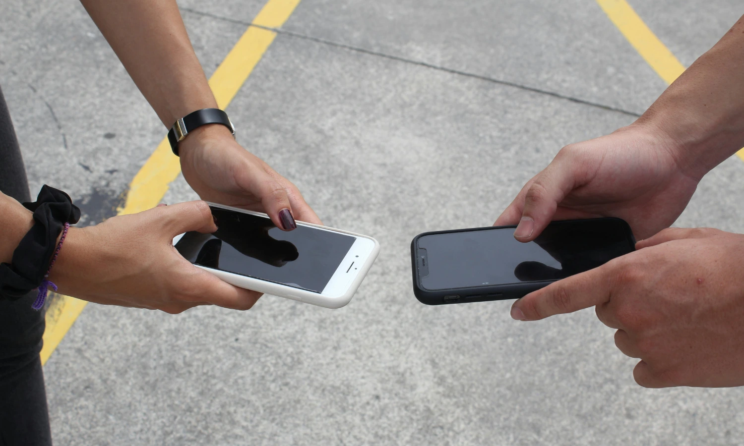 Two people hold smartphones toward each other, as if exchanging or showing their screens, on a concrete surface with yellow lines in the background.