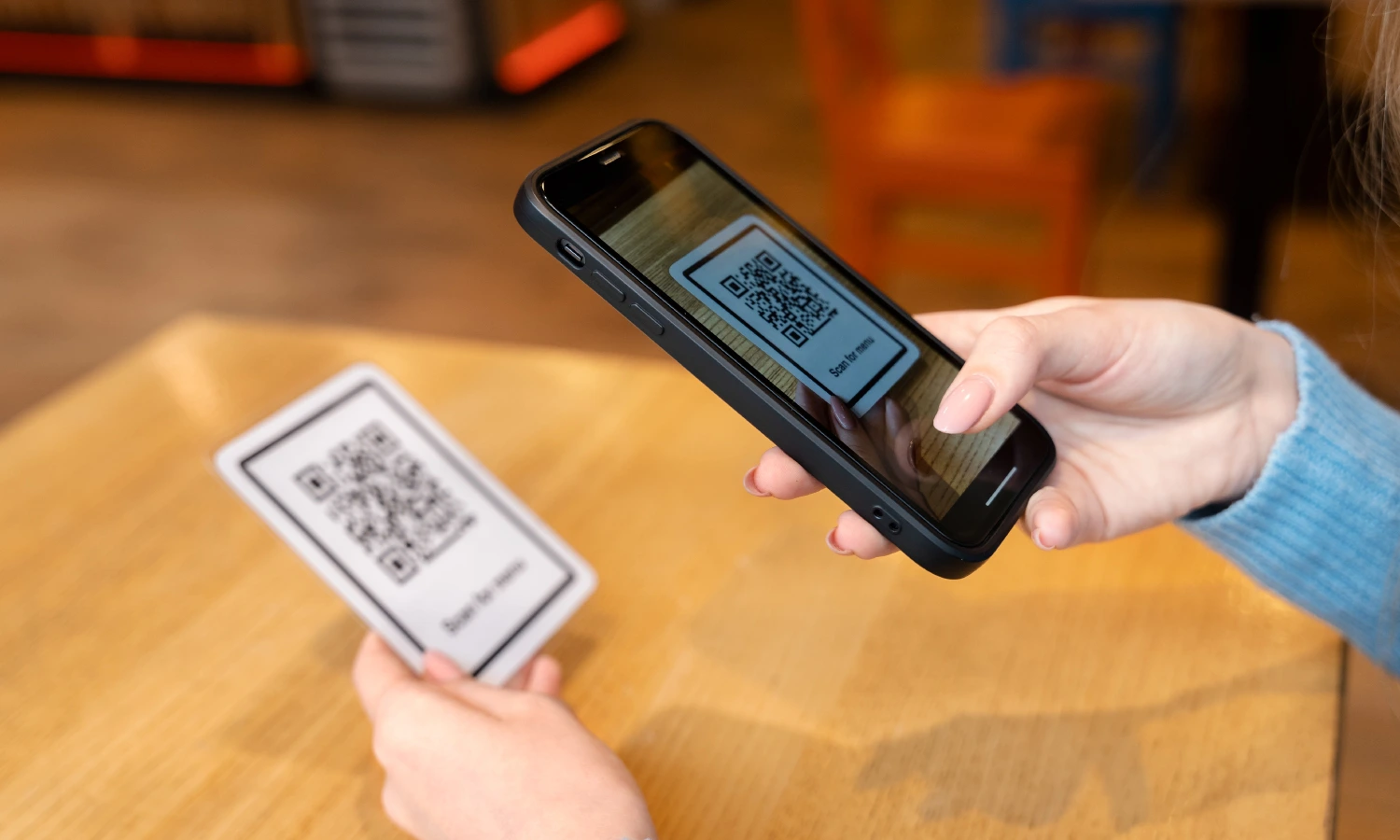 Person holds a QR code card while scanning it with a smartphone over a wooden table.
