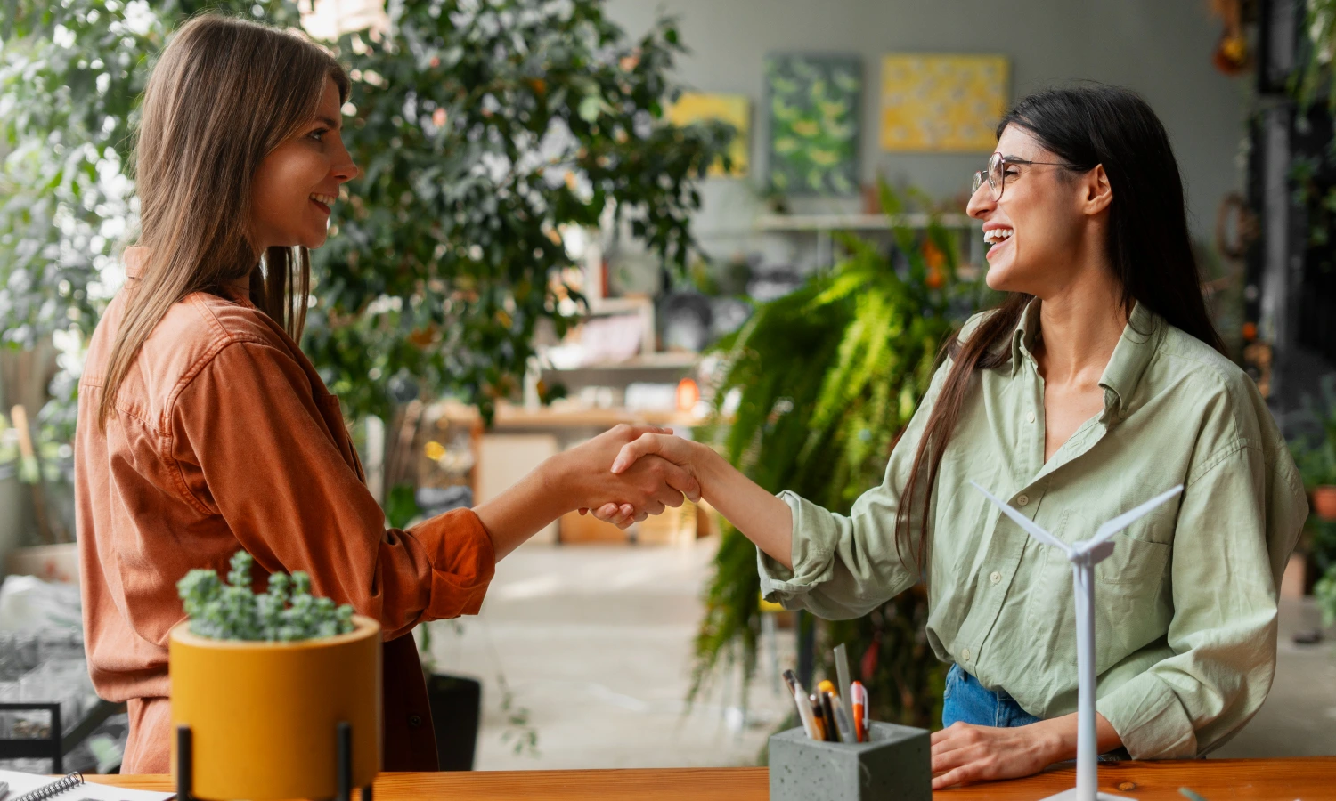Two women greet each other with a smile while shaking hands in a bright plant-filled space.