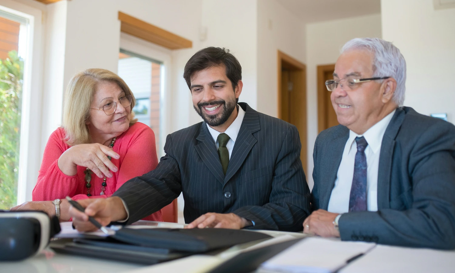 Three professionals collaborate at a well-lit table: a younger man in a suit smiles as he points with a pen, flanked by an older woman in pink and an older man in a gray suit.