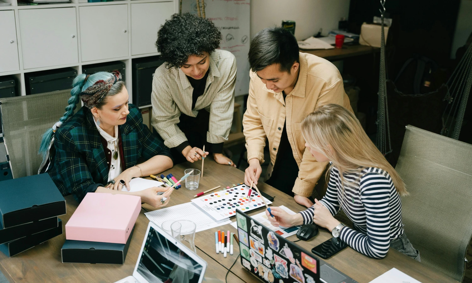 Four colleagues collaborate around a table, comparing color swatches and notes with laptops and markers on a design project.