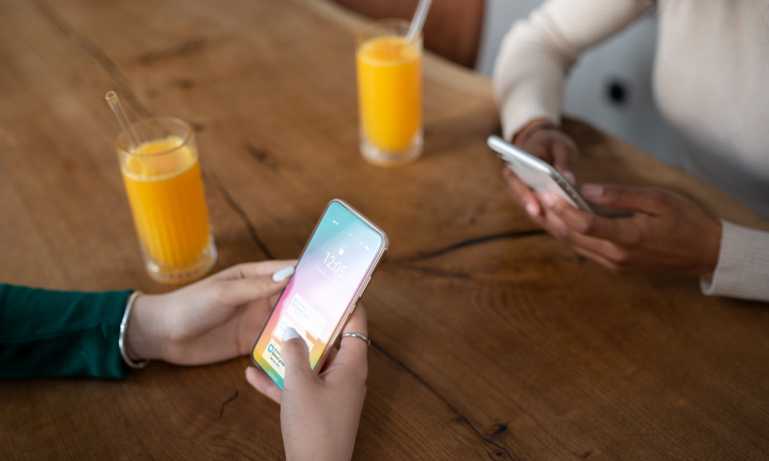 Two people sit at a wooden table, each using a smartphone; two glasses of orange juice are on the table behind them.