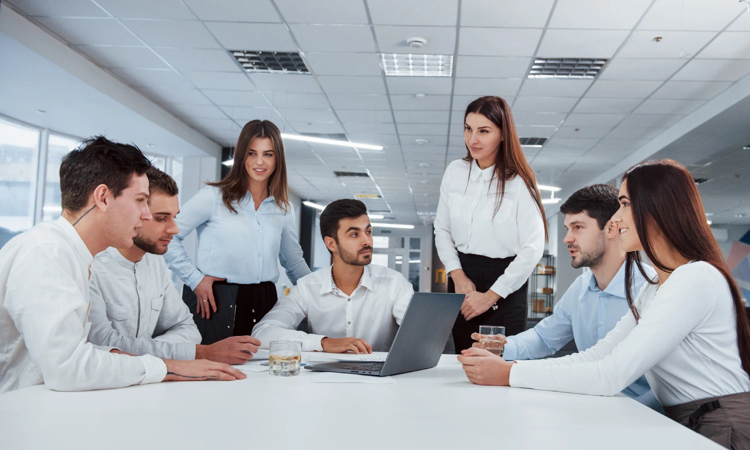 A diverse group of professionals around a conference table, reviewing a laptop screen in a modern office.